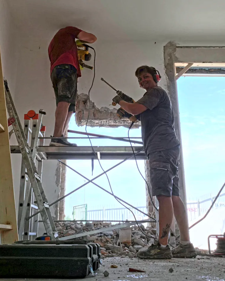 Anna e Salvatore al lavoro in un cantiere: Salvatore sul trabattello con il martello demolitore, Anna sorridente con il trapano e le cuffie